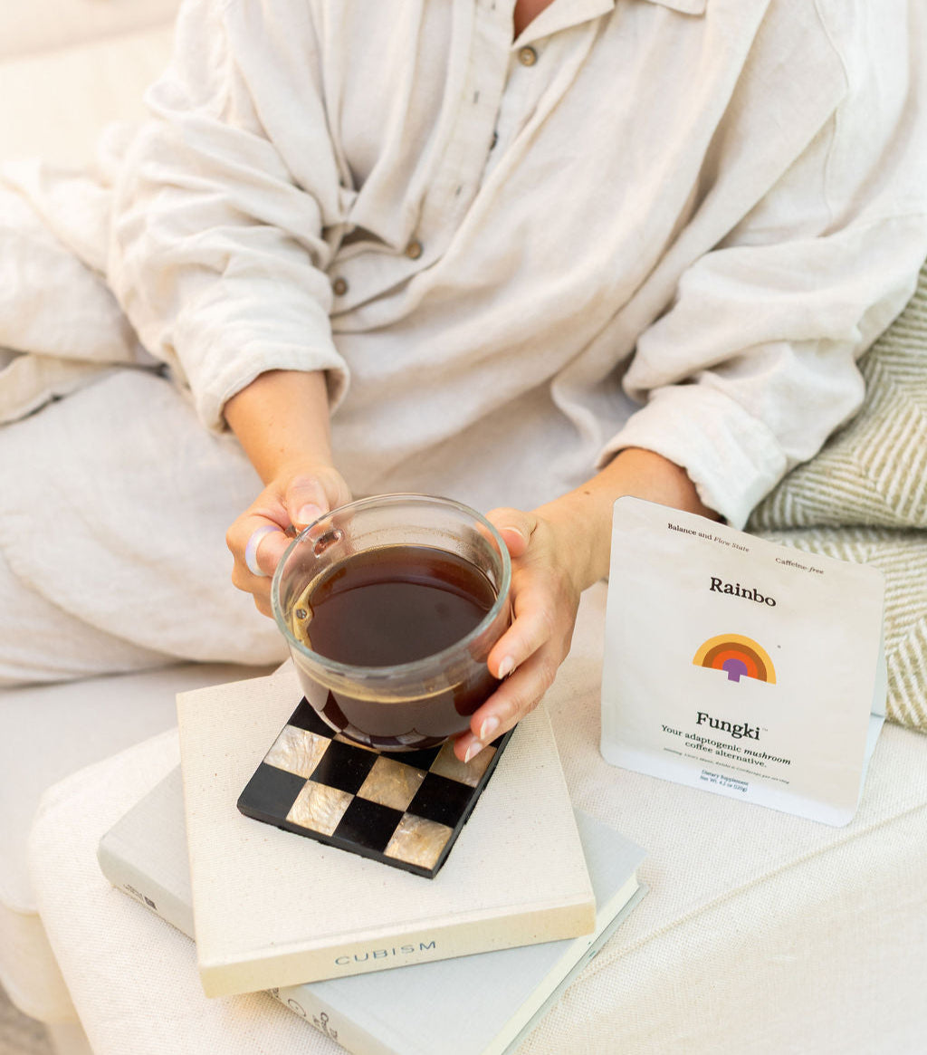 Person holding a mug of Fungki with a package of Rainbo Fungki on a checkered coaster.