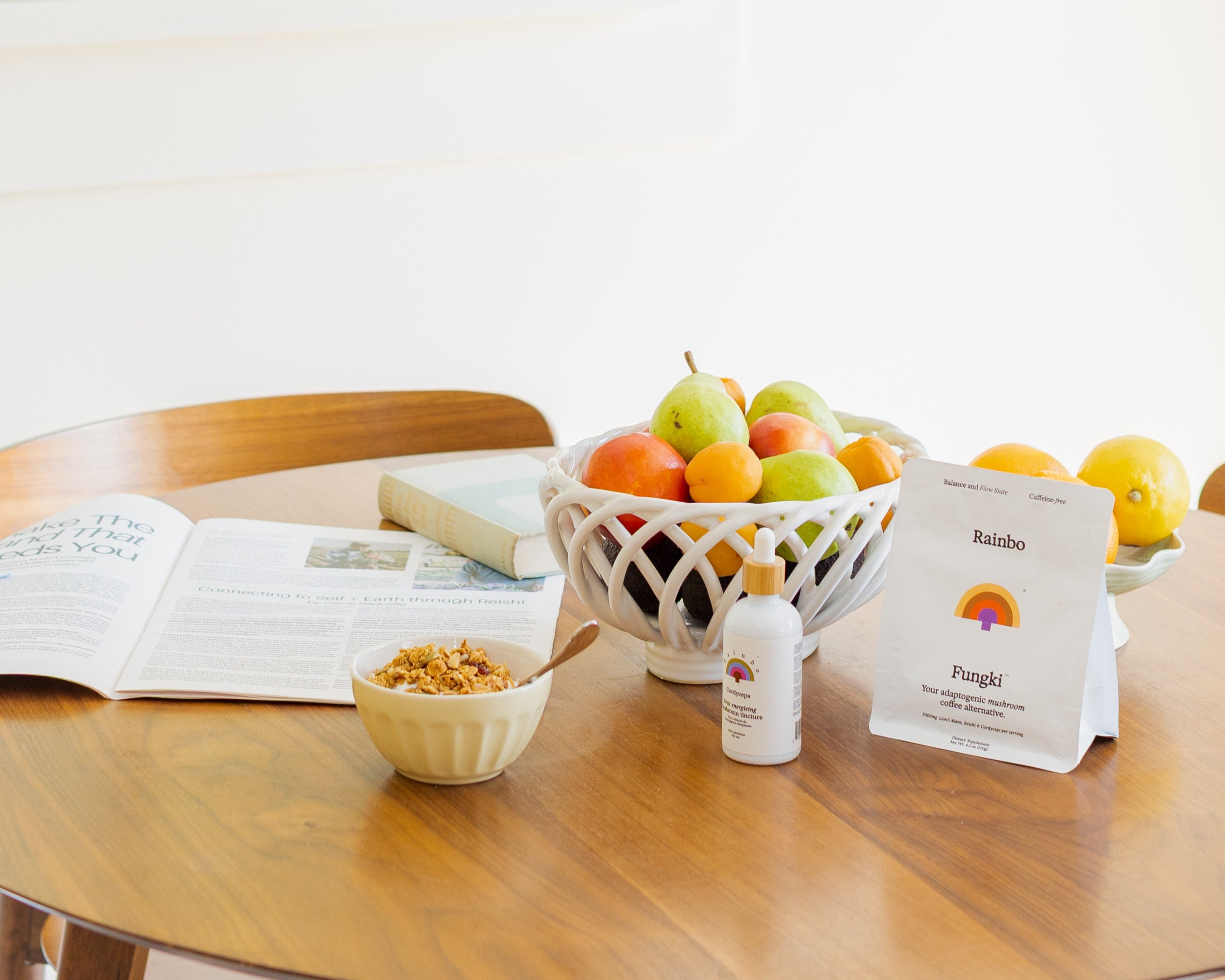 Table with a bowl of fruit, a Rainbo Cordyceps tincture bottle, a bowl of yogurt, a bag of Fungki mushroom coffee alternative and an open newspaper on a white background.