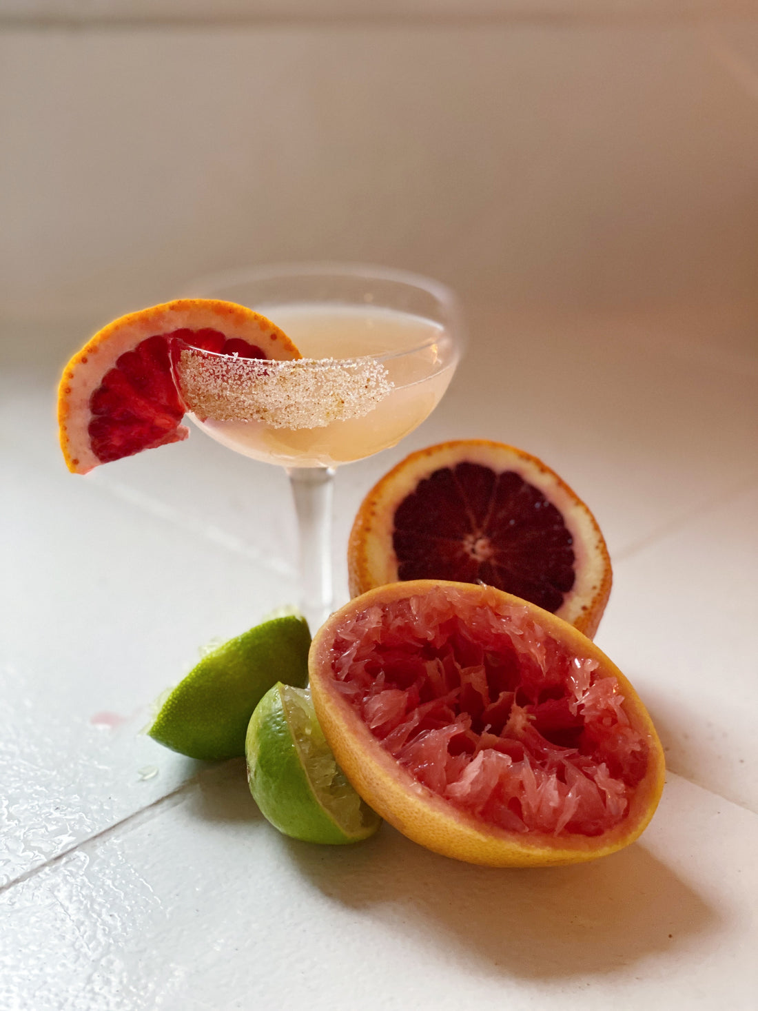 a coup cocktail glass is filled with a peachy liquid and a sugar rim. There is a sliced grapefruit as garnish on the glass and at the base of the glass is 2 squeezed limes, a squeezed grapefruit and half of a blood orange