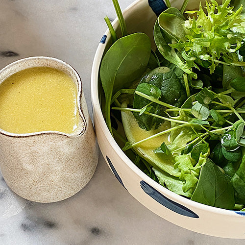half a bowl of salad is in view and on the other side of it is a pitcher filled with salad dressing. The salad dressing is a bright yellow colour and the salad itself is just green