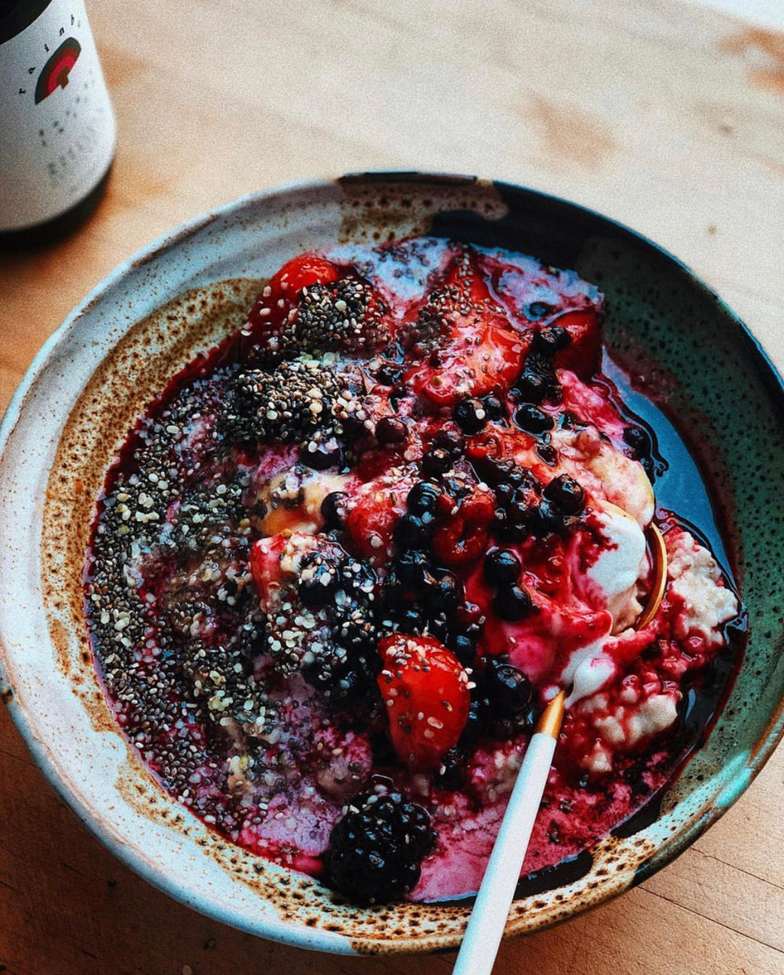 a bowl on a wooden counter with an oatmeal meal inside topped with chia seeds, blackberries, red strawberries, dark small blueberries, white coconut yogurt, and berry juices overflowing on the sides of the oatmeal. there is a spoon resting in the bowl