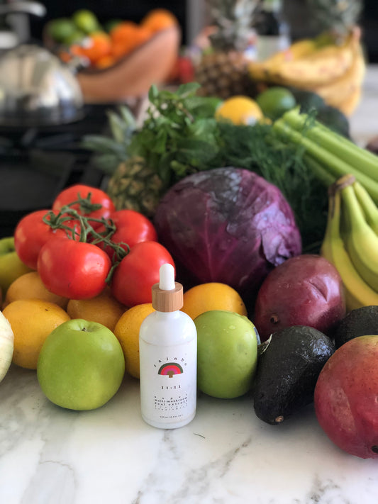 A white dropper bottle of Rainbo medicinal mushroom supplement on a counter filled with fresh colourful produce including apples tomatoes cabbage bananas avocado and greens
