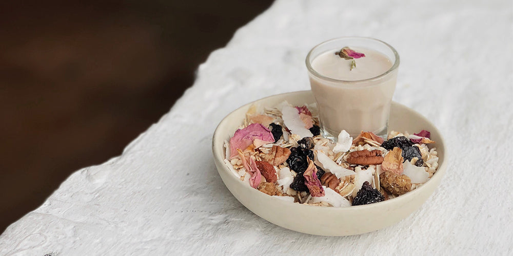 a white bowl sits on a white table with the dark wooden floor in the background. inside the bowl is artisanal granola made with colours pink, brown, white, dark blue pieces. Inside the bowl is also a short clear glass of nut milk made with mushrooms