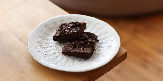 a white ceramic plate sits on the corner of a counter top made of light coloured wood. on top of the plate sits two dark brown chocolate brownies with flaked sea salt on top