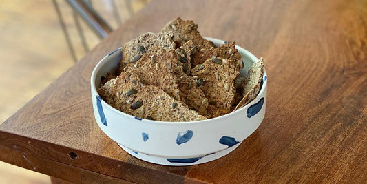 A white bowl with blue spots is on a dark wood table with crackers mounded inside. The crackers show evidence of varying seeds and nuts and are a light brown colour. 