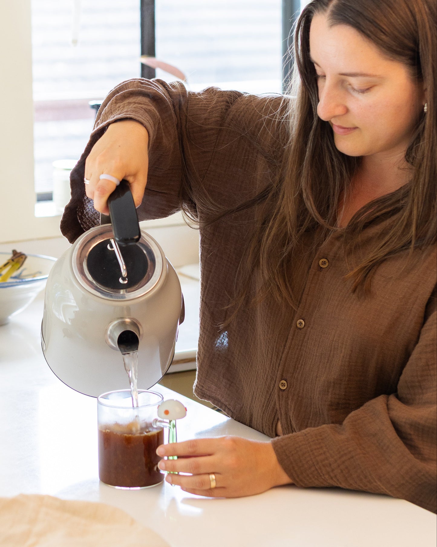 Woman pouring hot water from a kettle into a glass with Fungki on a kitchen counter.
